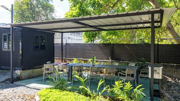 A covered outdoor patio with tables and chairs, surrounded by greenery and a fence.