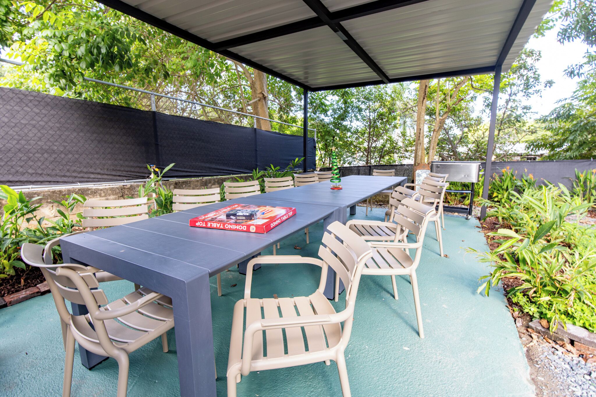 Un patio techado al aire libre con una mesa larga y sillas, rodeado de follaje y una cerca negra.