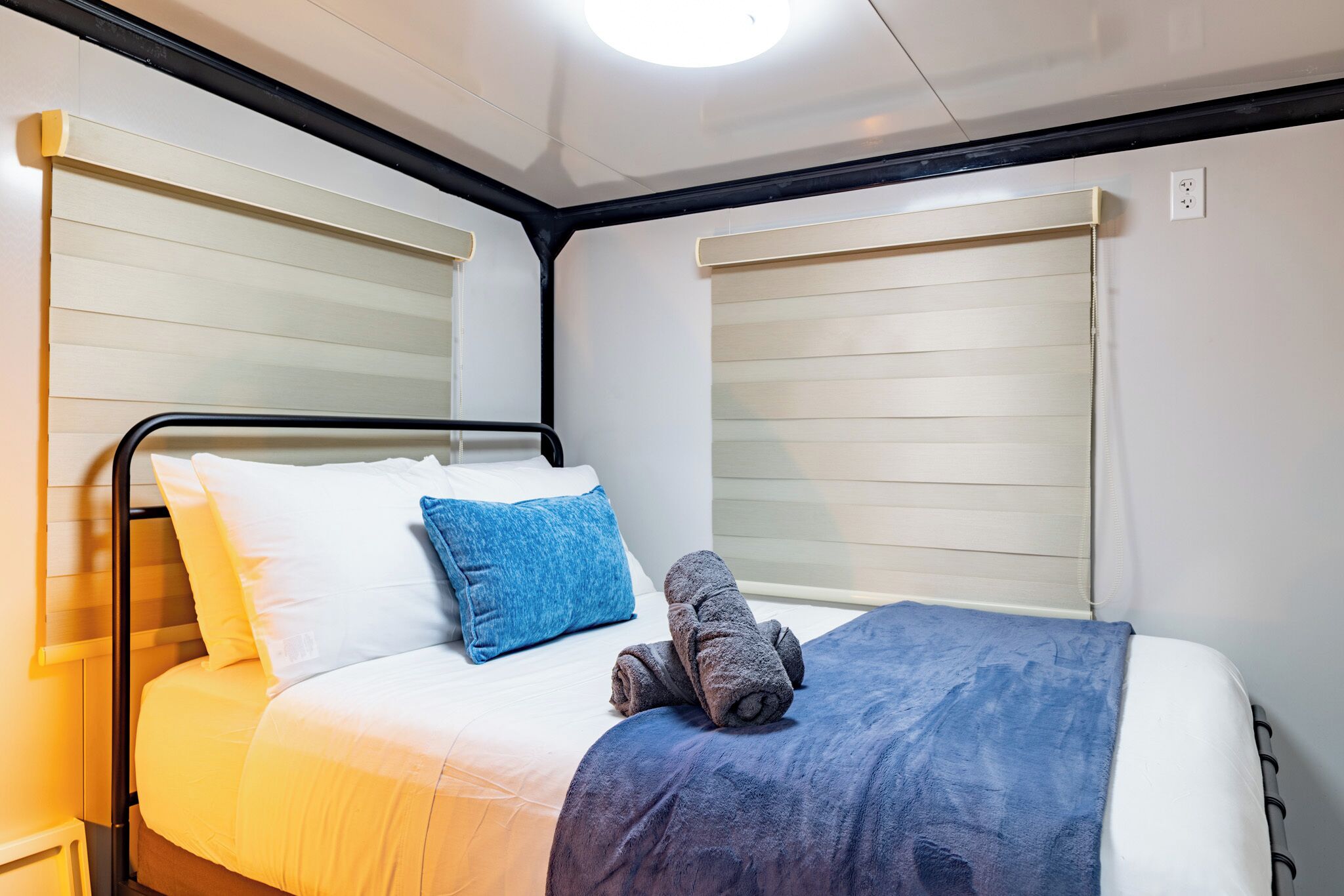 A neatly made bed with a blue blanket and a rolled towel, white and blue pillows, and a black metal frame.
