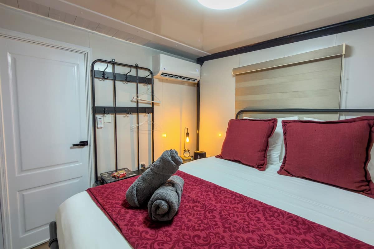 A hotel room with a bed, a red patterned bedspread, a white door, a coat rack, and an air conditioning unit.