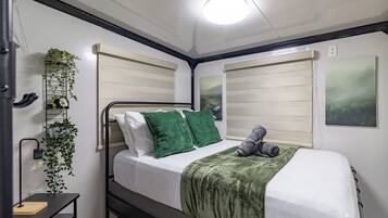 Un dormitorio ordenado con cama, almohadas verdes, manta gris, mesita de noche con lámpara y una planta en la pared.