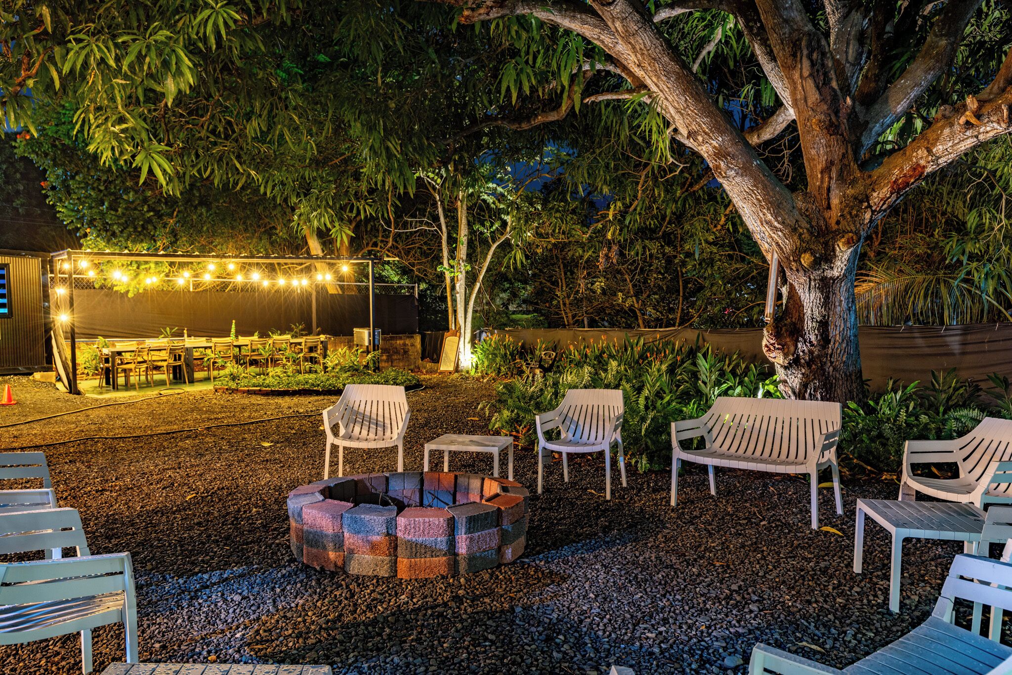 Un espace extérieur aménagé avec des chaises blanches et un foyer, entouré d’arbres, et une zone de repas couverte illuminée de guirlandes lumineuses.