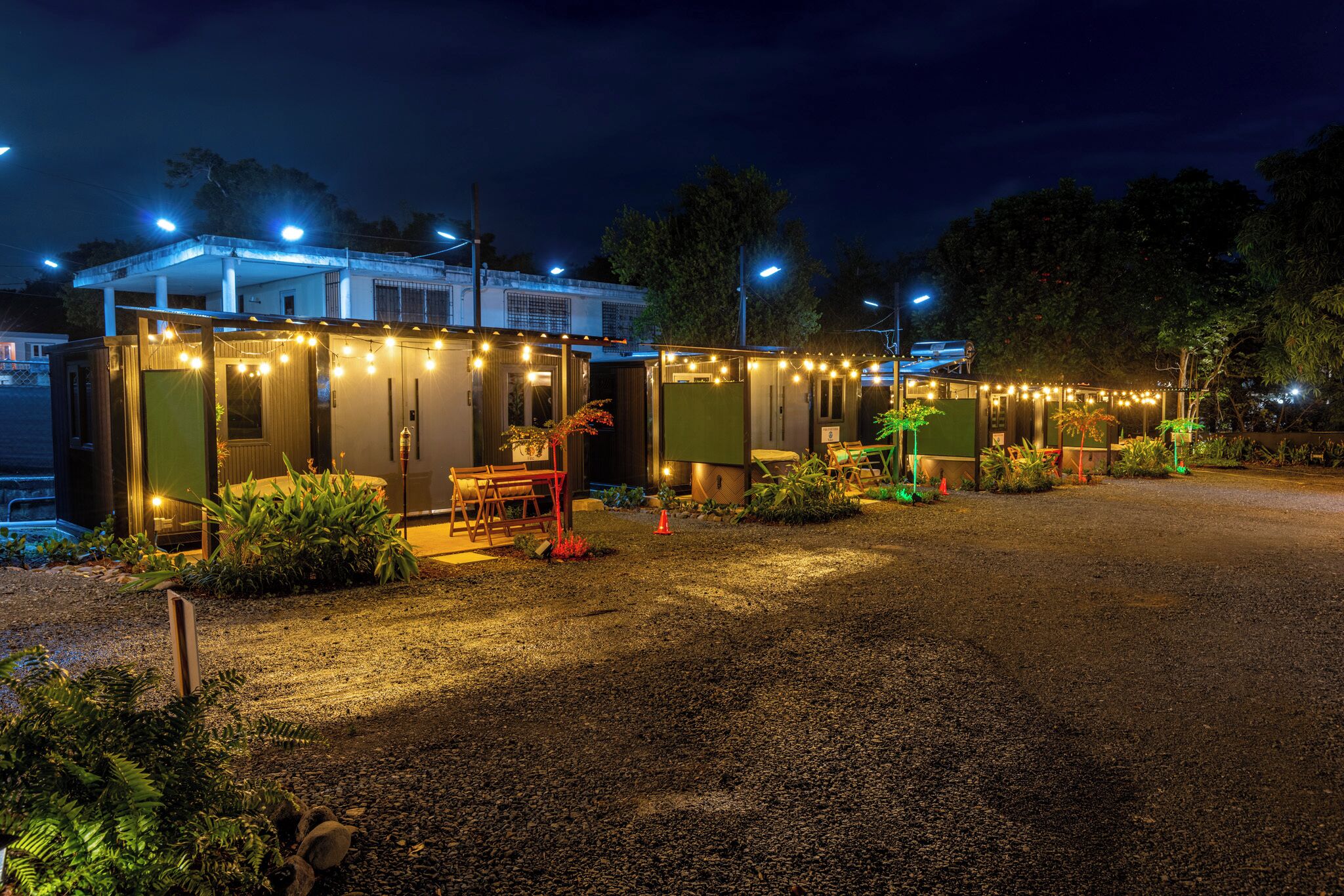 A row of small, single-story cabins with string lights, surrounded by gravel and greenery.