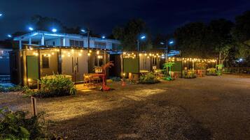 A row of small, single-story cabins with string lights, surrounded by gravel and greenery.