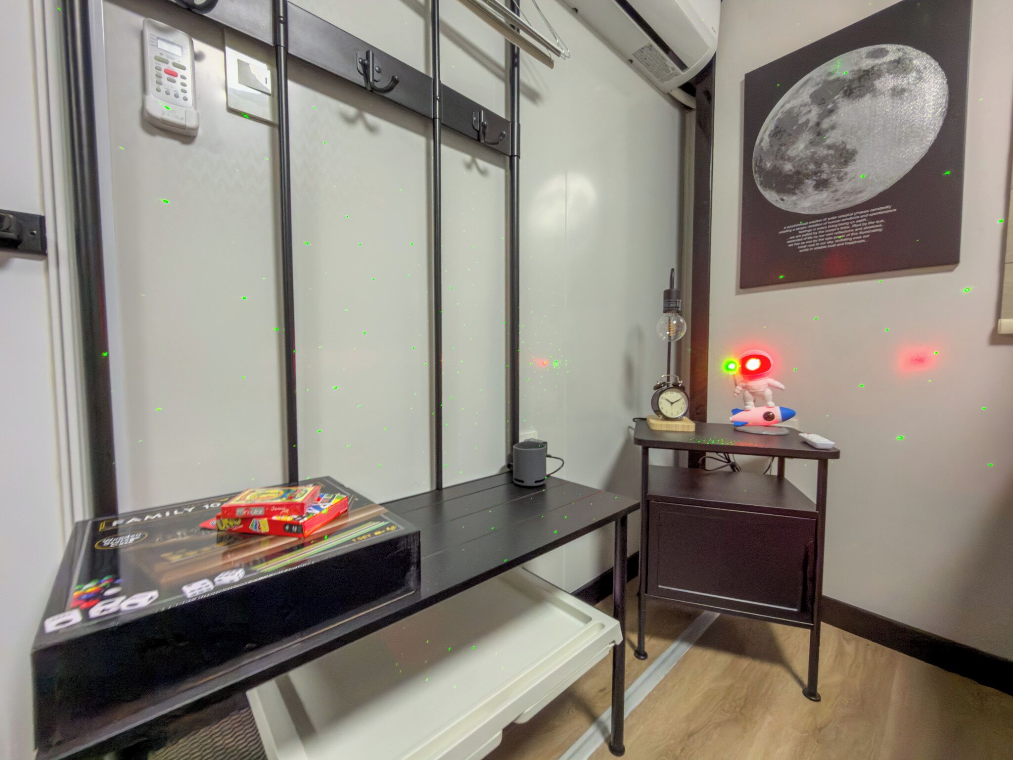 A hotel room with a desk, a small table with a clock and a toy, a wall-mounted phone, and a poster of the moon.