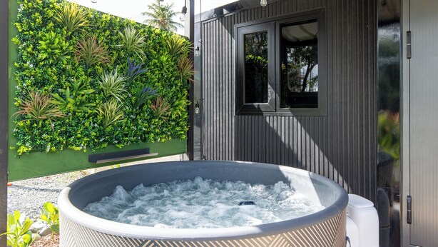Ein Whirlpool in einem überdachten Außenbereich mit einer Holzterrasse, umgeben von üppiger Vegetation und einem Holzhaus mit einem Fenster.