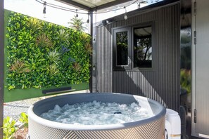 A hot tub in a covered outdoor area with a wooden deck, surrounded by lush greenery and a wooden building with a window. - Tatloanis Village (Vega Baja)
