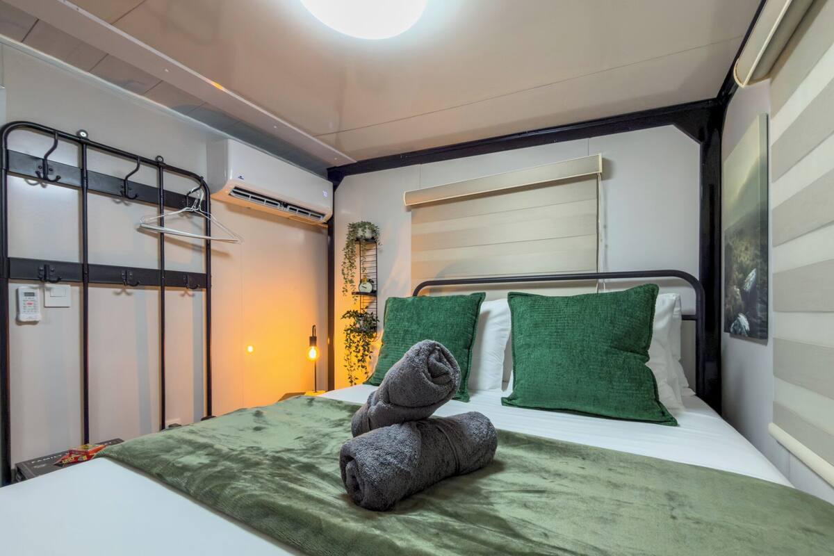 A hotel room with a bed, green pillows, a green blanket, a wall-mounted air conditioner, and a window with blinds.