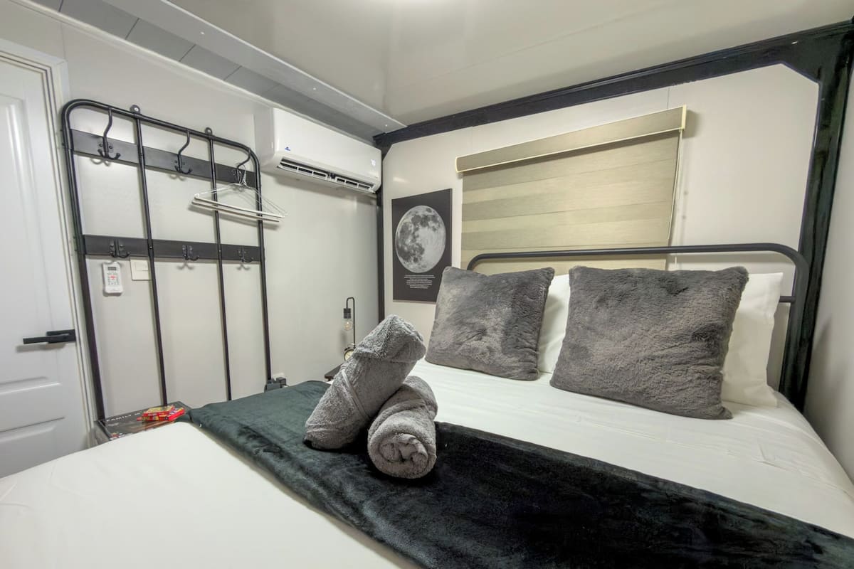 A compact hotel room with a bed, a black and white headboard, a wall-mounted air conditioner, a coat rack, and a framed picture on the wall.