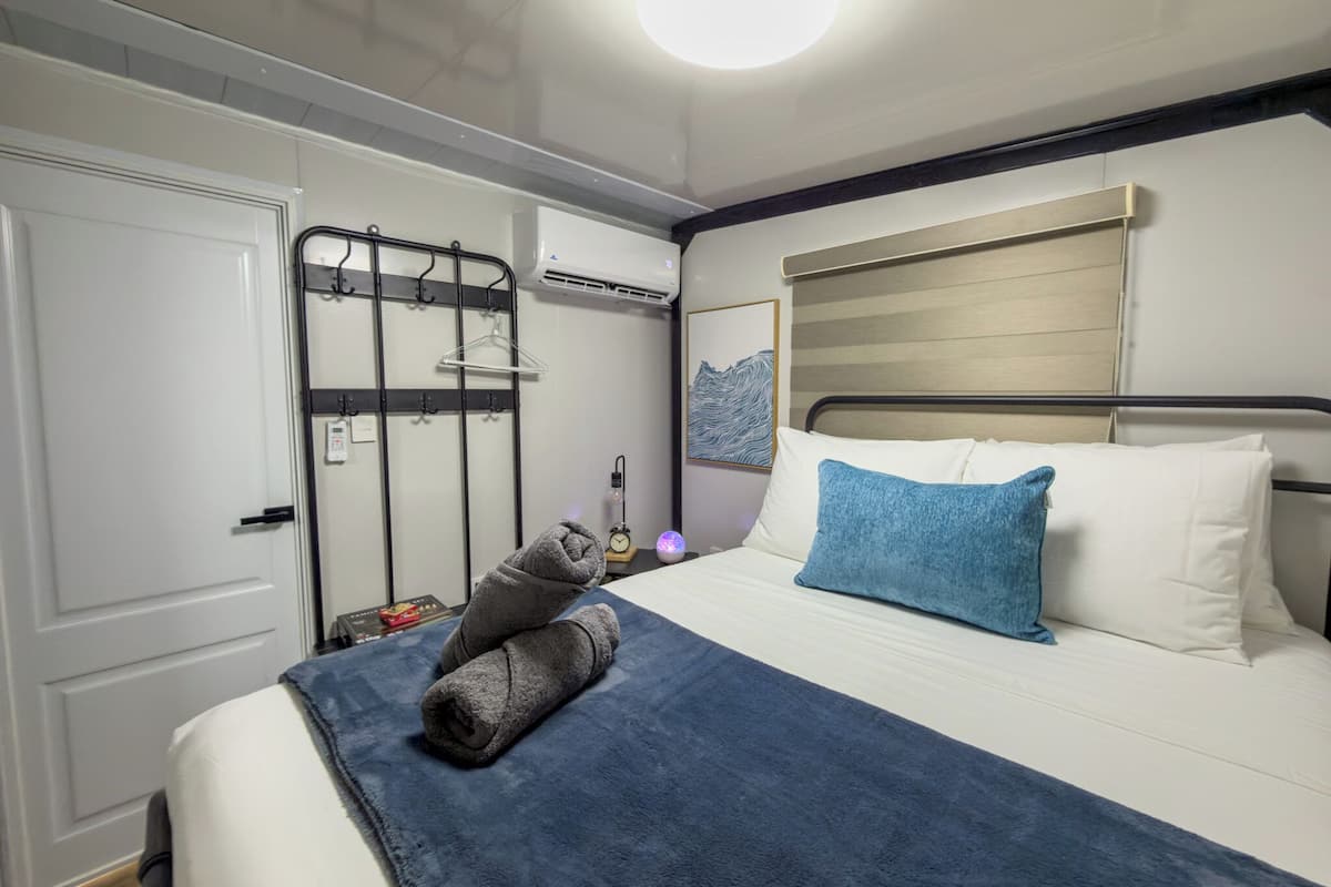 A neatly made bed with a blue blanket and pillows, a bedside table with a lamp, and a wall-mounted air conditioner.