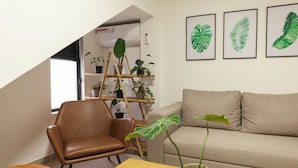 Ein Wohnzimmer mit braunem Ledersessel, beige-farbenem Sofa, Couchtisch aus Holz und einem Blumentopf. An der Wand befinden sich gerahmte Blätterbilder und ein Bambusregal mit Pflanzen.