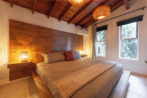 A bedroom with a wooden headboard, a large bed with multiple pillows, bedside lamps, and a window with a view of trees. - Cozy Astorga Home (Medellín)