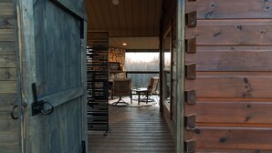 A wooden cabin interior with a wooden door, a stone fireplace, wicker furniture, and a view through the door.