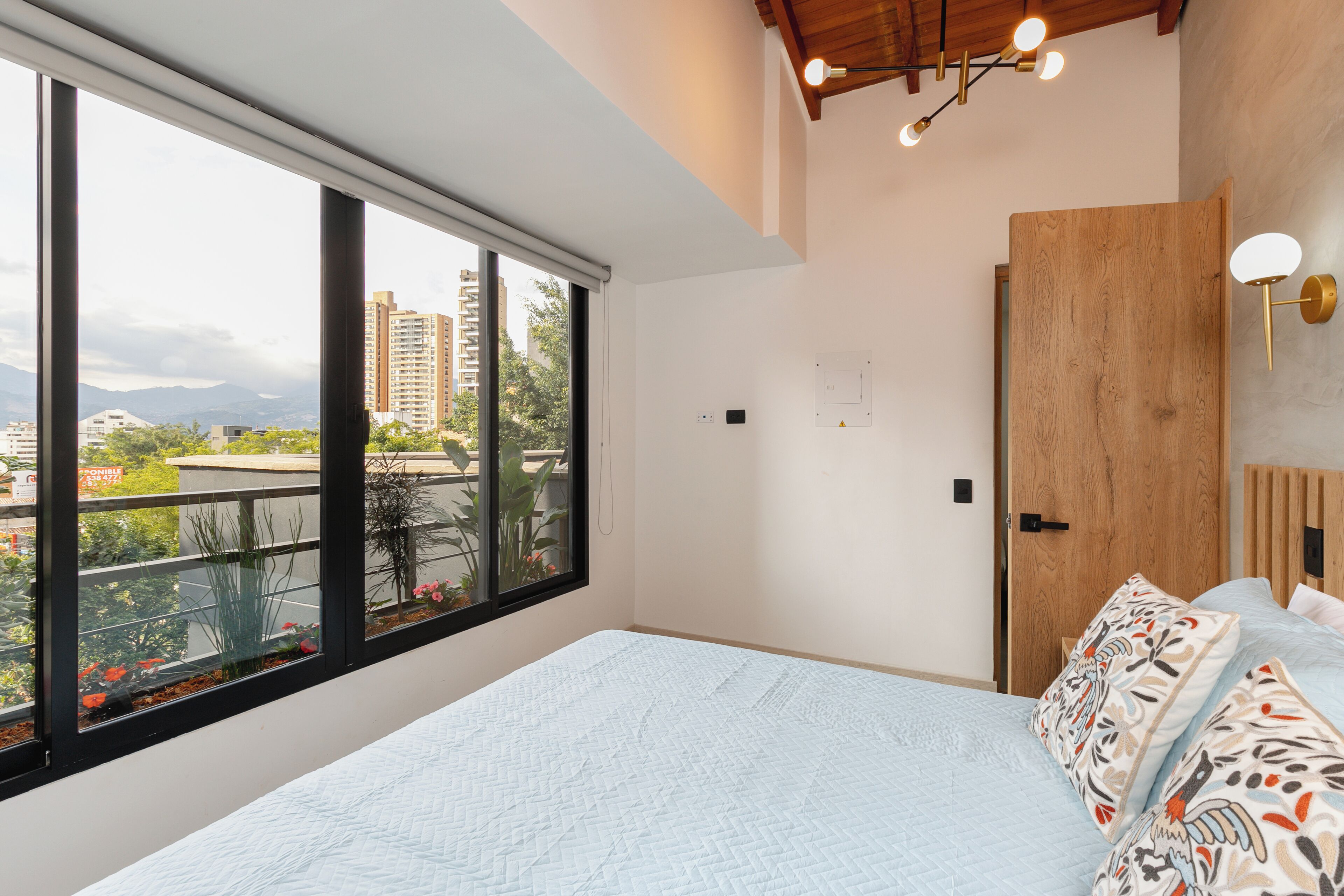 Un dormitorio con un ventanal grande, una cama con almohadones estampados, una puerta de madera y una lámpara en el techo.