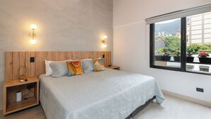 Ein Schlafzimmer mit Bett, Nachttischen, Wandleuchten und einem Fenster mit Blick auf Grünflächen und Gebäude.
