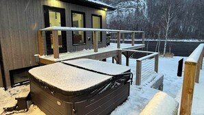 Une cabane en bois dotée d’un jacuzzi recouvert de neige, d’une terrasse et offrant une vue sur un paysage enneigé.