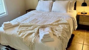 En pænt redt seng med hvide sengetæpper og et foldet håndklæde, en sengetabletlampe og et vindue med persienner.