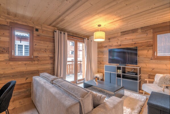 A wooden cabin interior with a sofa, TV, and sliding glass doors to a balcony. - Appartement Jorat - Welkeys (Megève)
