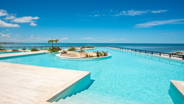 Ein klares, blaues Schwimmbecken mit Meerblick, umgeben von Palmen und einer Steindeckterrasse.