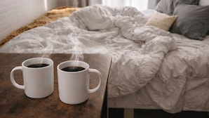 A bedroom with a bed, two white mugs on a table, and a window with curtains.