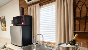 A kitchen with a built-in sink, a microwave, a refrigerator, a window with curtains, and a bottle of champagne on the counter.