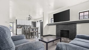 Ein modernes Wohnzimmer mit grauen Sofas, einem Kamin und einem Essbereich, im Hintergrund ist eine Küche zu sehen.