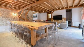 A spacious living area with a large wooden dining table surrounded by clear chairs, a stone wall, a staircase, a sofa, a TV, and a washing machine. - Le Gite du Relais - PARGUES (PARGUES)