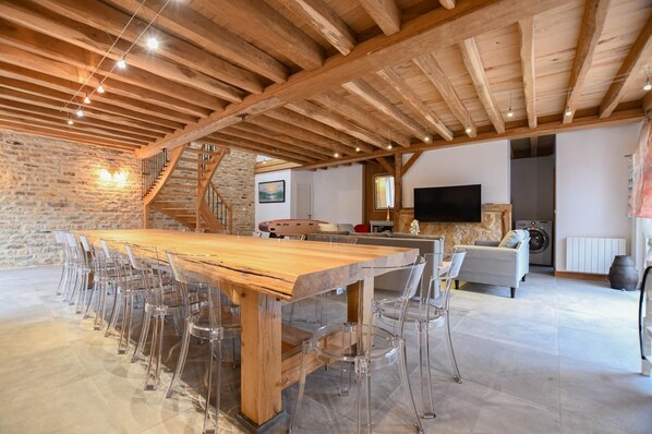 Amplia sala de estar con una gran mesa de comedor de madera rodeada de sillas transparentes, pared de piedra, escalera, sofá, televisor y lavadora.
