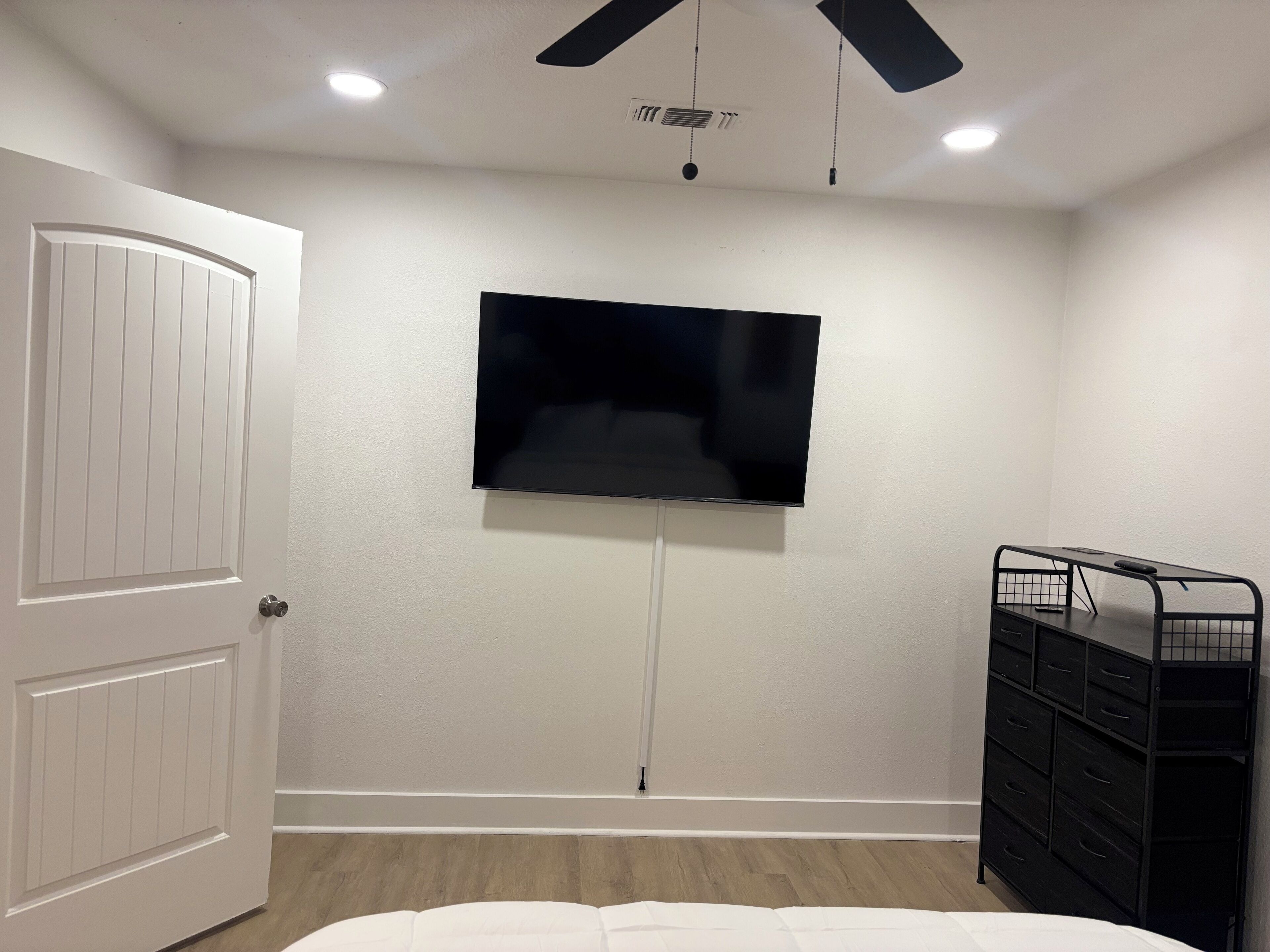 A bedroom with a bed, a TV mounted on the wall, a ceiling fan, and a dresser.