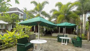 Un patio con bancos y mesas verdes bajo sombrillas, rodeado de palmeras y un edificio.