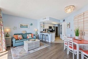 A modern living room with a sofa, coffee table, and a kitchen area with a dining table and chairs. - Roxanne Towers 1004 (Myrtle Beach)