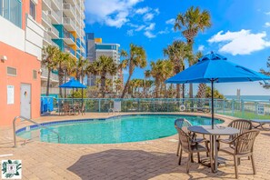 A swimming pool with lounge chairs and umbrellas, surrounded by palm trees and a view of the ocean. - Roxanne Towers 1004 (Myrtle Beach)