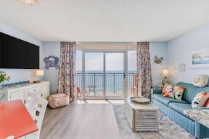 A living room with a sofa, a table, a TV, and a balcony with a view. - Roxanne Towers 1004 (Myrtle Beach)