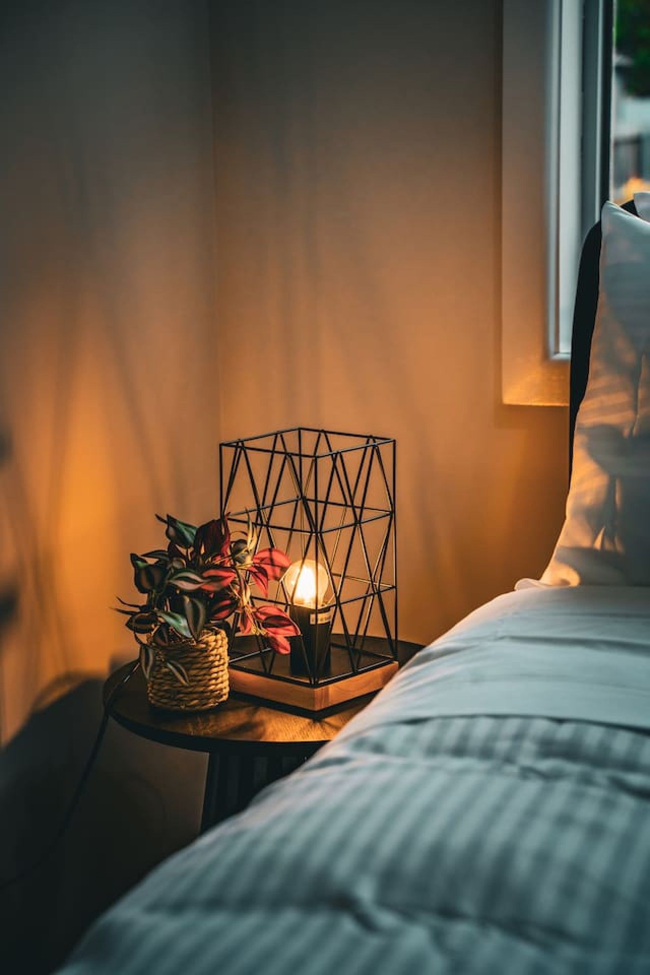A bed with a headboard, a bedside table with a geometric lamp, and a basket of plants.