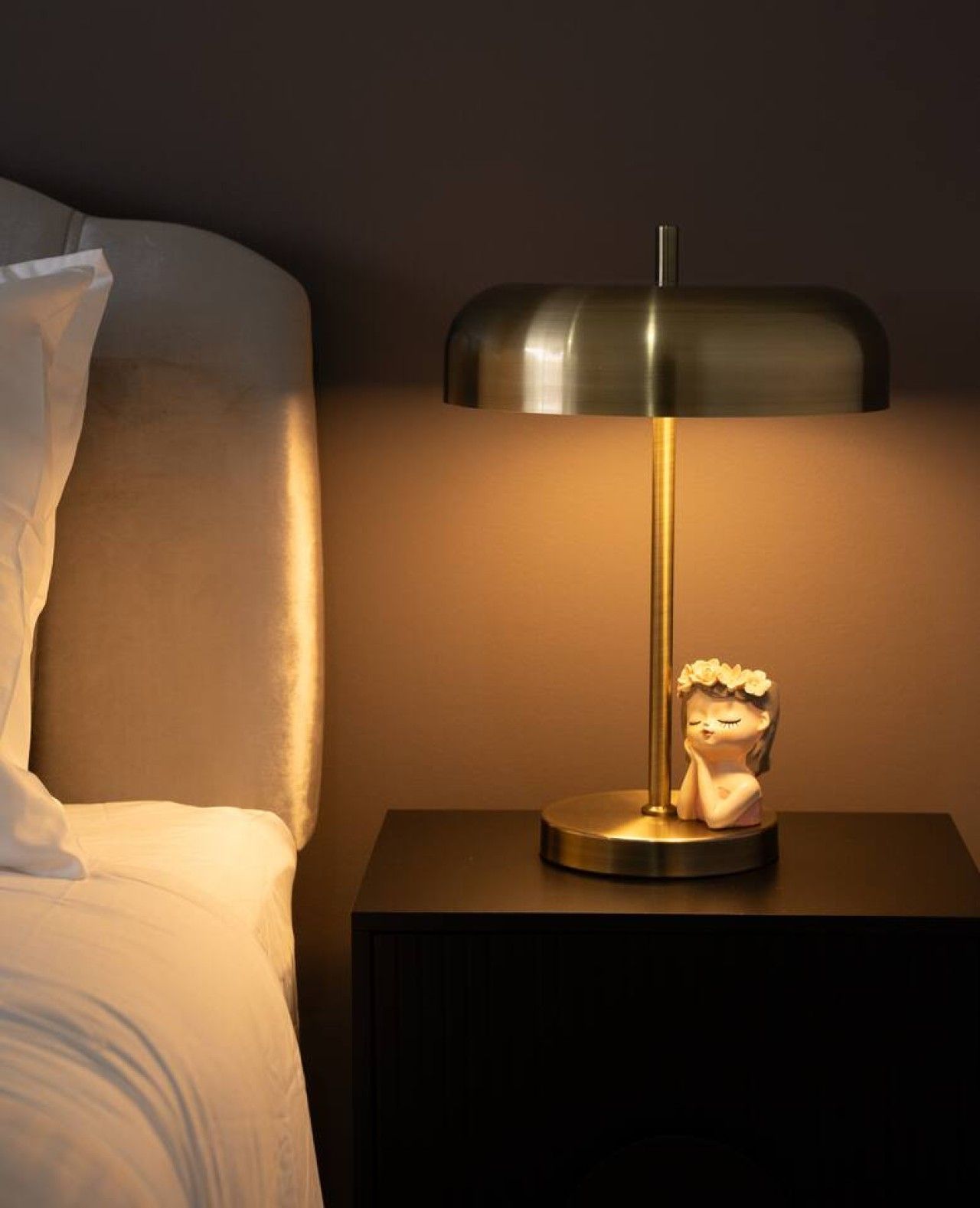 A bedside table with a lamp and a figurine, next to a bed with white bedding.