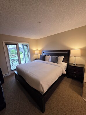 A bedroom with a large bed, two bedside tables with lamps, a window with blinds, and a carpeted floor.