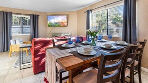 A dining area with a table set for four, a red sofa, and a TV mounted on the wall. - Point Loma Hideaway · Relaxing Hot Tub + AC (San Diego)