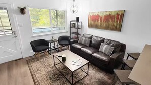 Ein Wohnzimmer mit einer Ledersofa, zwei schwarzen Sesseln, einem Couchtisch und einem Teppich. An der Wand hängt ein Gemälde, und ein Bücherregal ist mit Büchern gefüllt.