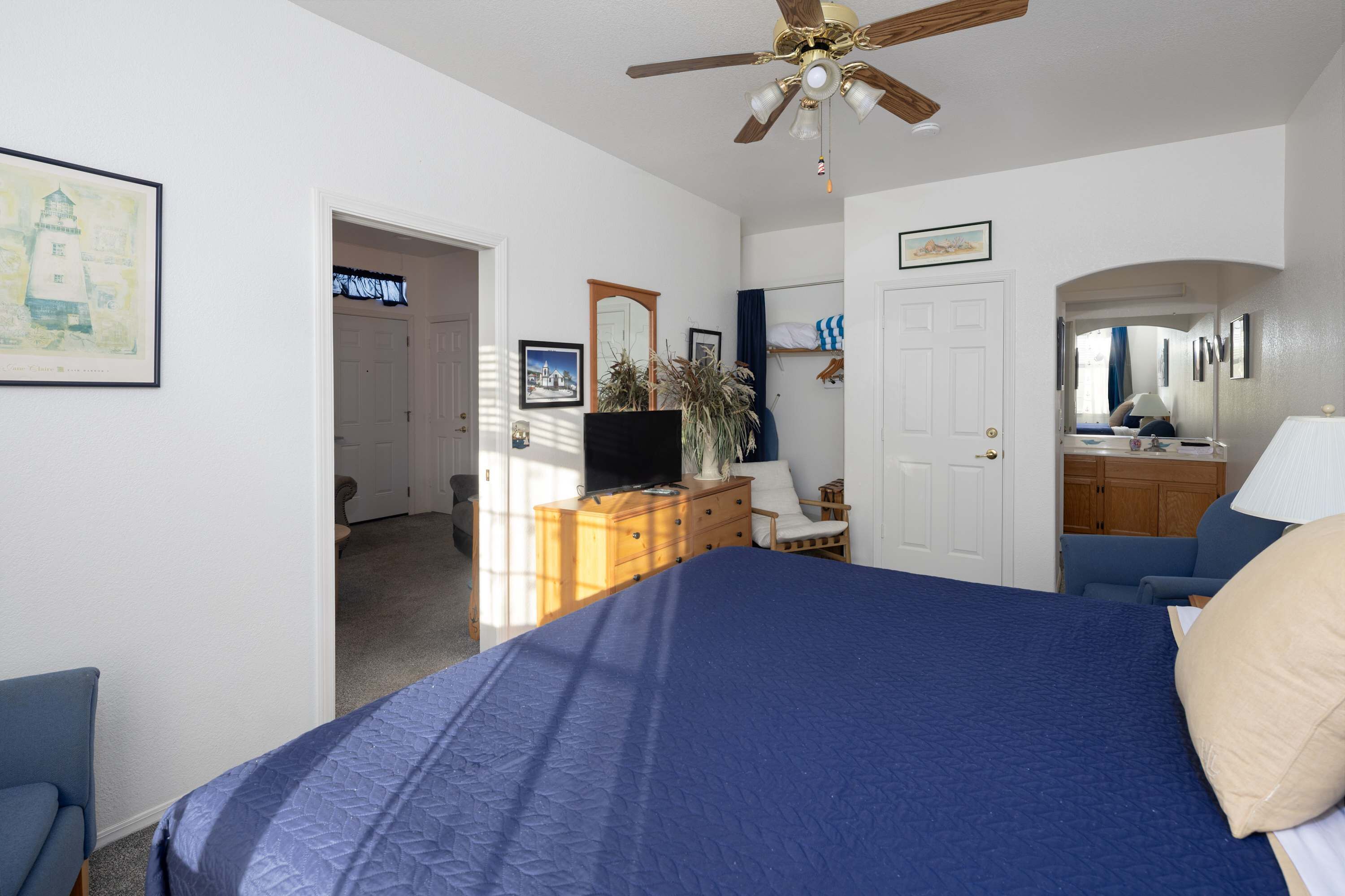 Une chambre à coucher comprenant un lit, un ventilateur de plafond, une commode, un téléviseur, une chaise et un bureau avec une lampe.