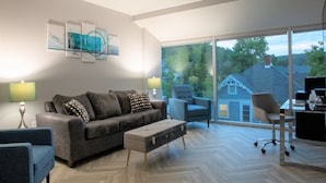 A modern living room with a grey sofa, blue armchair, and a glass coffee table. There is a large window offering a view of a house and trees. - Cozy 2-bedroom hotel suites just two blocks from Downtown Athens. (Athens)