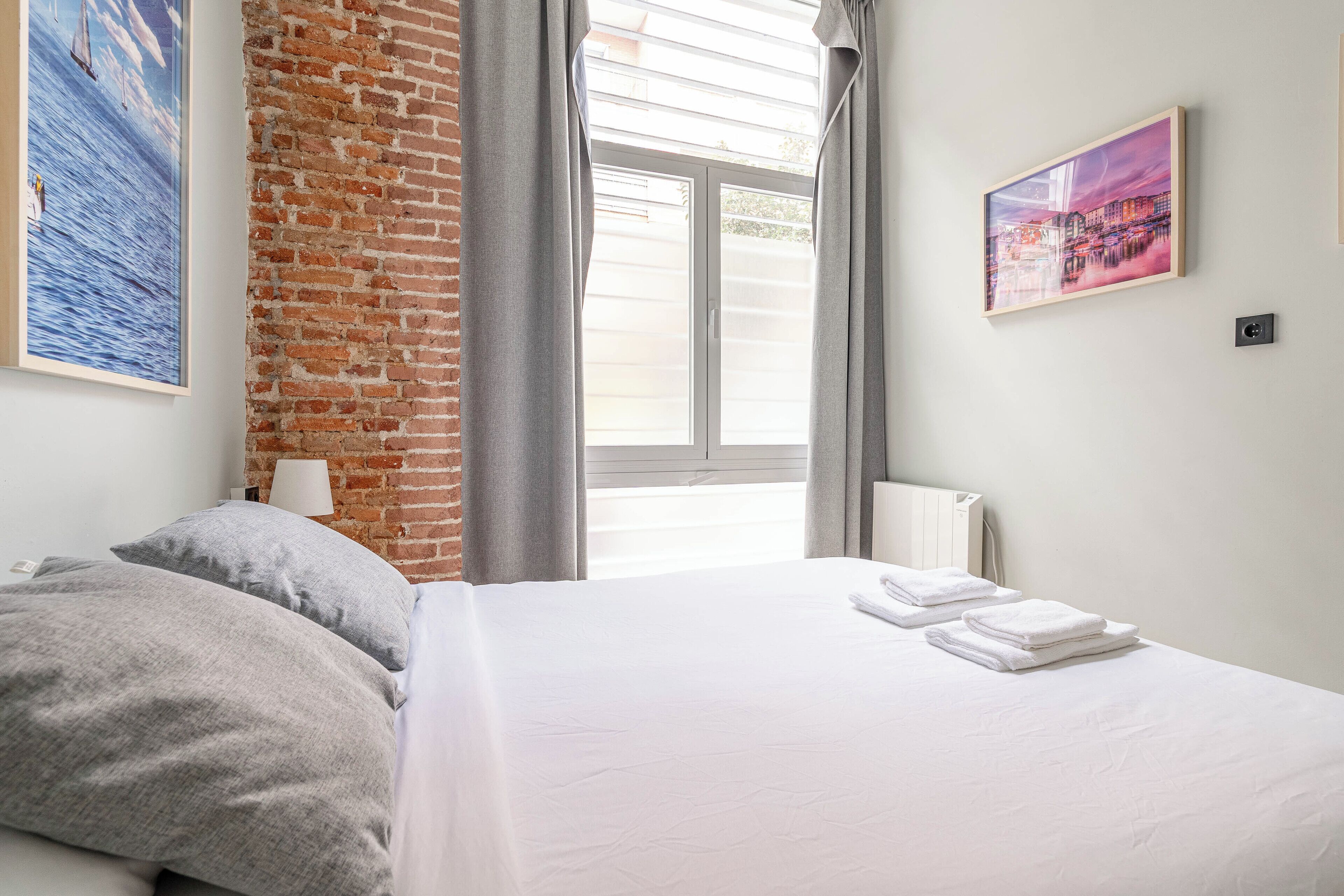 Hotellihuone, jossa on tiiliseinä, suuri ikkuna, jossa on verhot, valkoisilla lakanoiden peitetty sänky ja kaksi taulua seinillä.