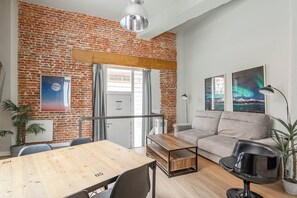 Ein Zimmer mit Backsteinwand, einem Holztisch, einer Couch und zwei Blumentöpfen.