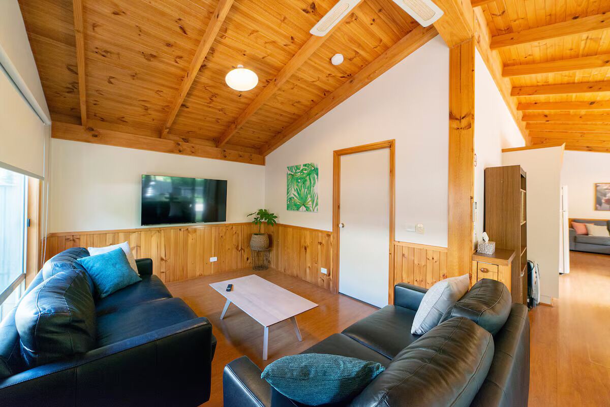 A spacious living room with wooden ceilings and walls, a flat-screen TV mounted on the wall, a sofa, a coffee table, and a cabinet.