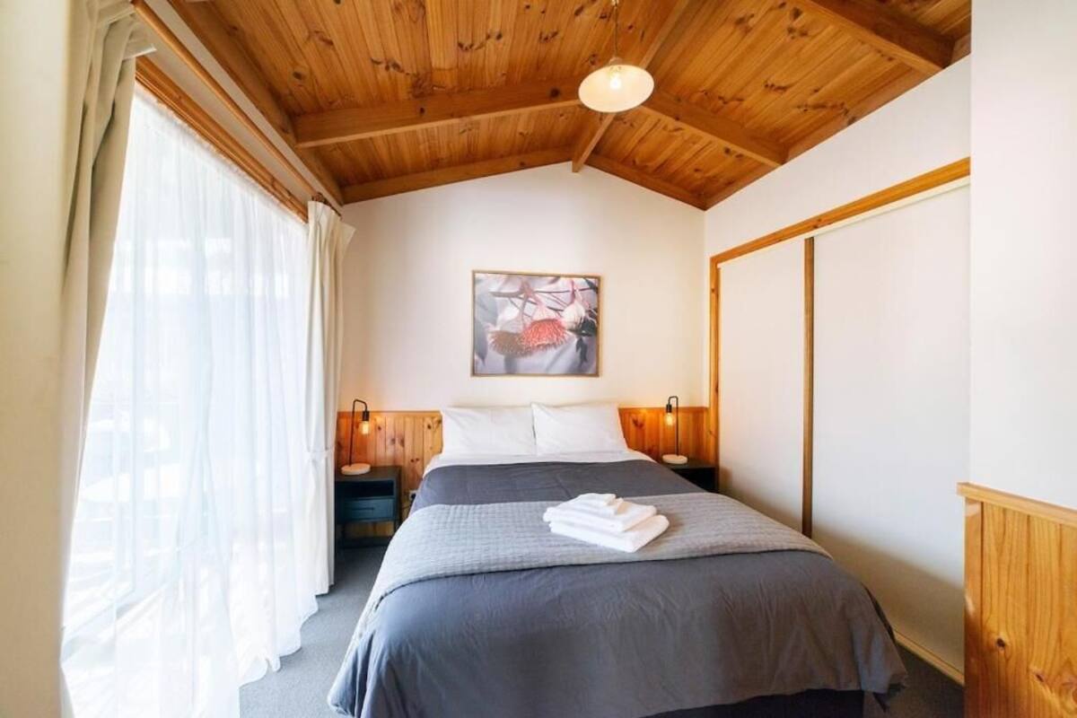 A bedroom with a wooden ceiling, a bed with a grey bedspread, white pillows, a framed picture on the wall, and two bedside lamps.