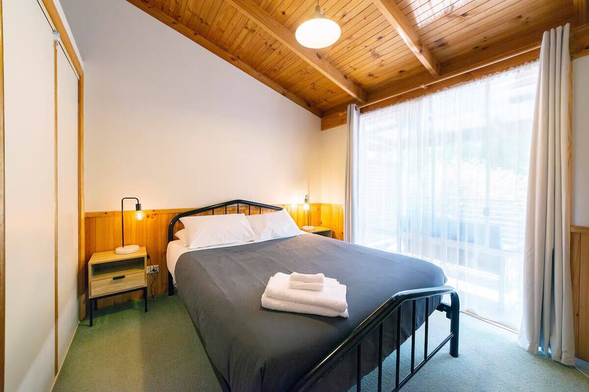 A single bed with a black frame, a wooden headboard, and a grey bedspread. There are two bedside tables with lamps, a window with sheer curtains, and a wooden ceiling.