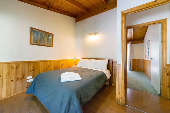 Un dormitorio con techo de madera, una cama con colcha gris, una almohada blanca y una toalla doblada sobre la cama. Hay un cuadro en la pared y una lámpara sobre la cama.