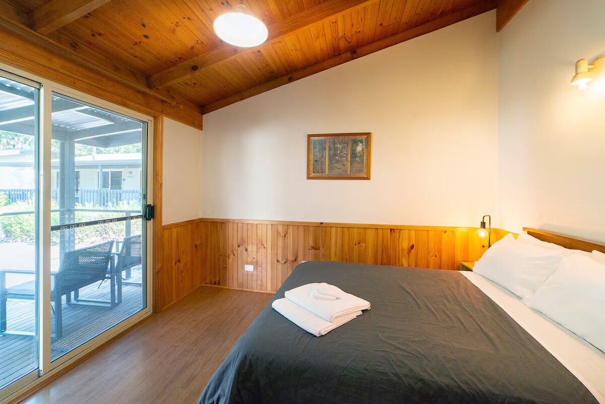 Een slaapkamer met een houten plafond, een bed met witte lakens, een ingelijst schilderij aan de muur, en een schuifdeur naar een buitenruimte met een tafel en stoelen.