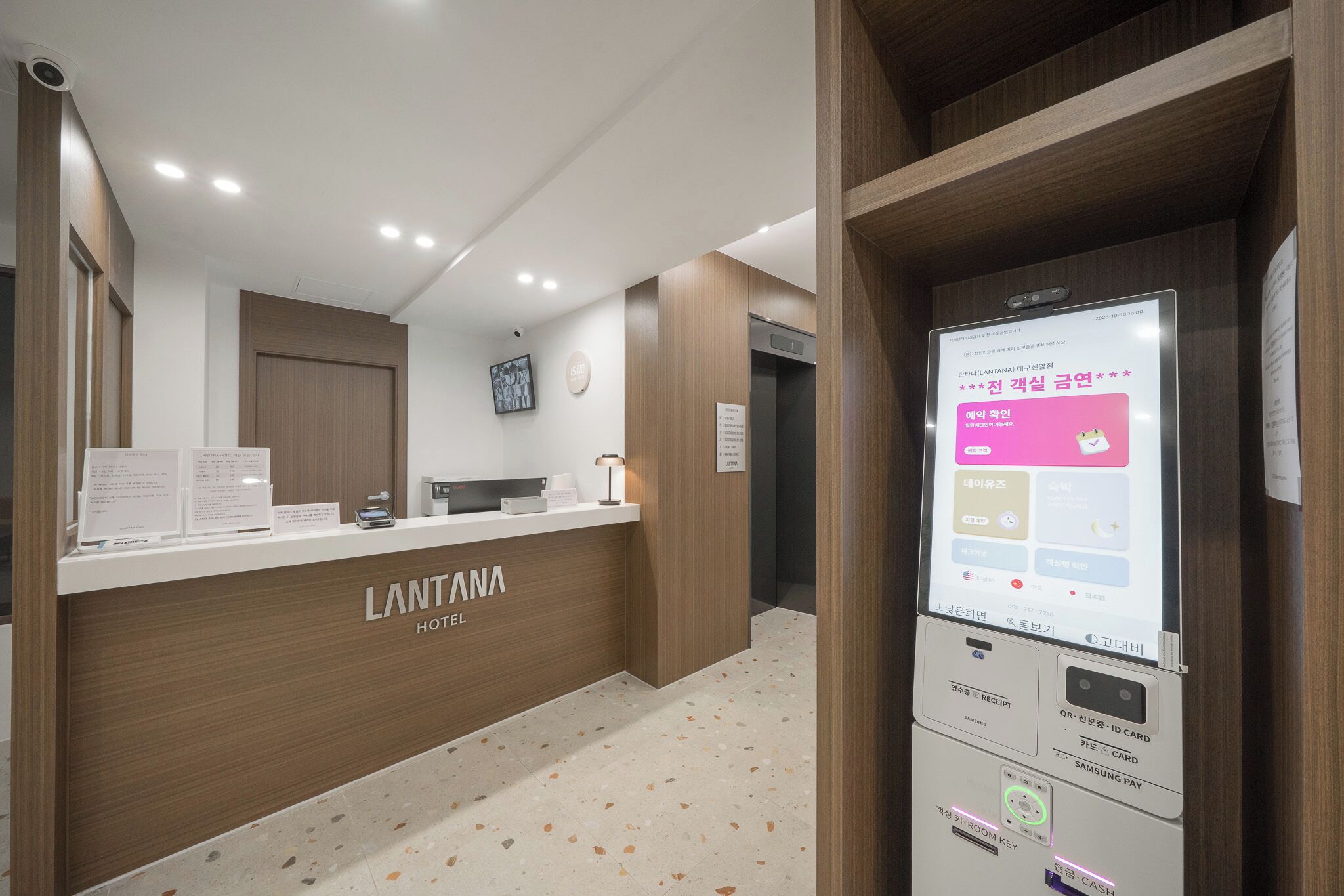 Ein Hotel-Empfangsbereich mit einem Holzbüro, einer digitalen Informationskiosk und einer Wanduhr.