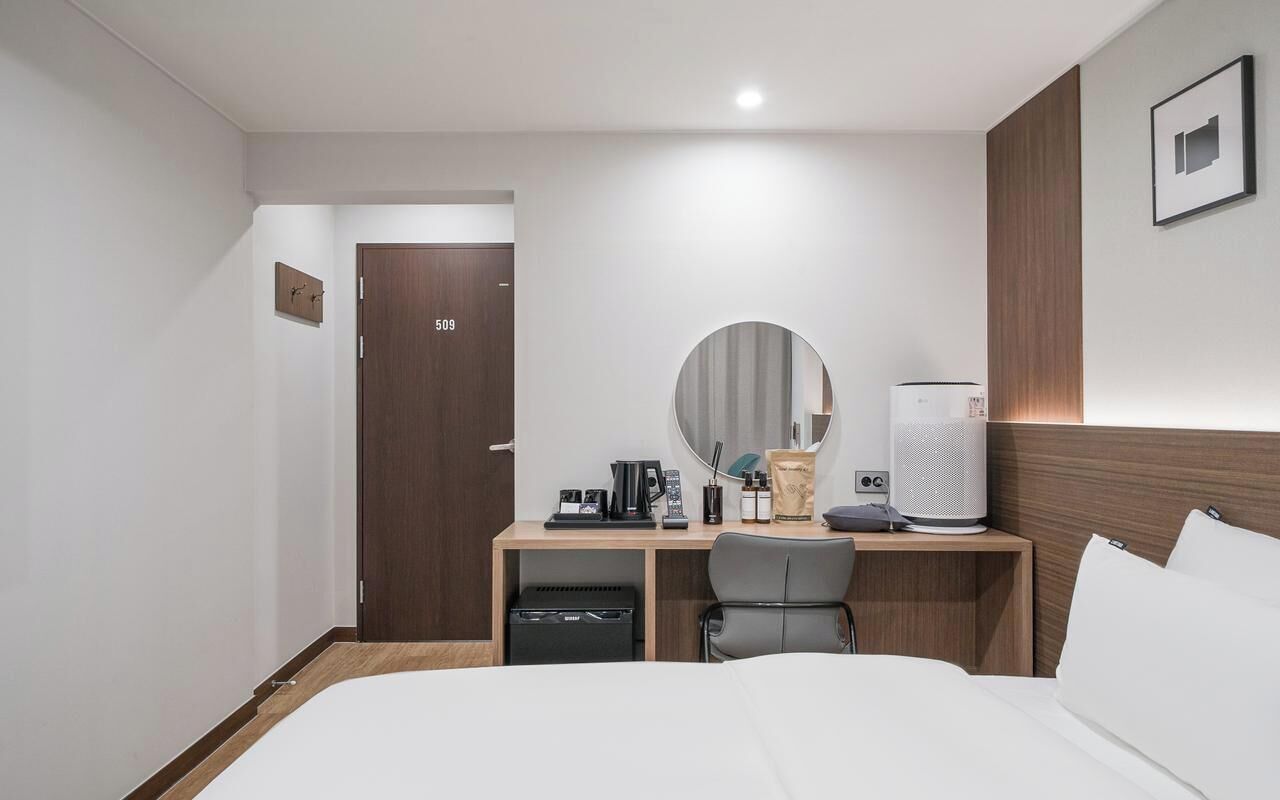A hotel room with a bed, a desk with a computer, a chair, a coffee maker, a speaker, and a door.