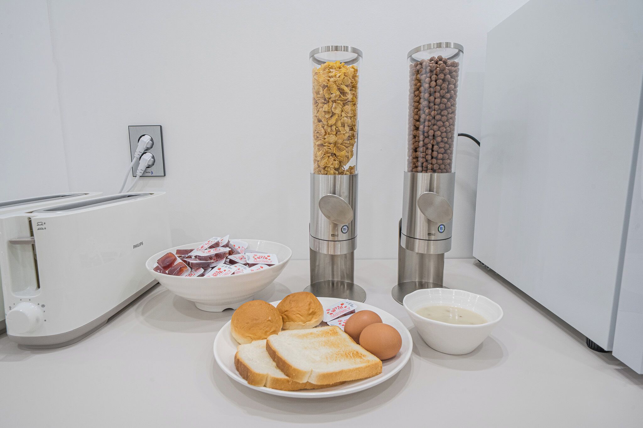 A kitchen with a toaster, two food dispensers, a bowl of cereal, a plate with bread and eggs, and a bowl of milk.